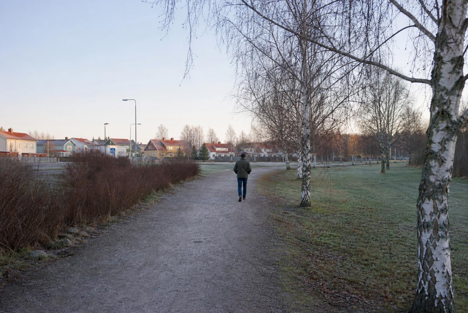En person går på en grusväg genom ett ljust svenskt villaområde i morgonljus, sedd bakifrån.